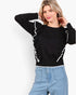 A woman modeling a black long-sleeve knit sweater with white ruffled trim along the shoulders and sides, paired with light blue jeans against a plain white studio background.