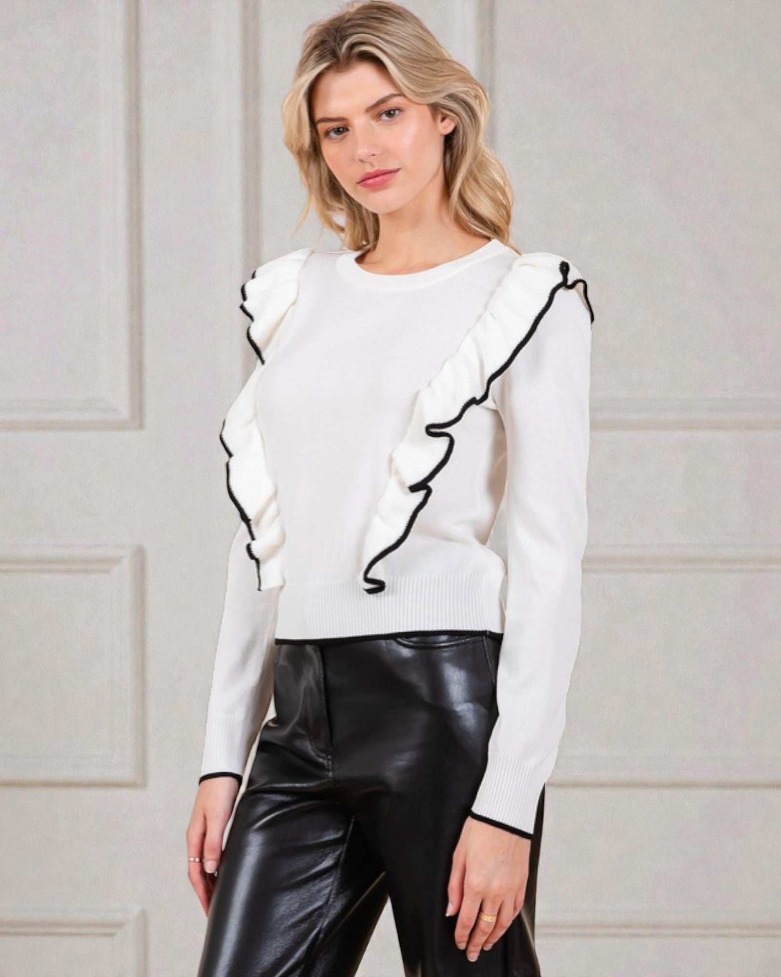 Woman wearing a white sweater with ruffled details and black pants against a light background