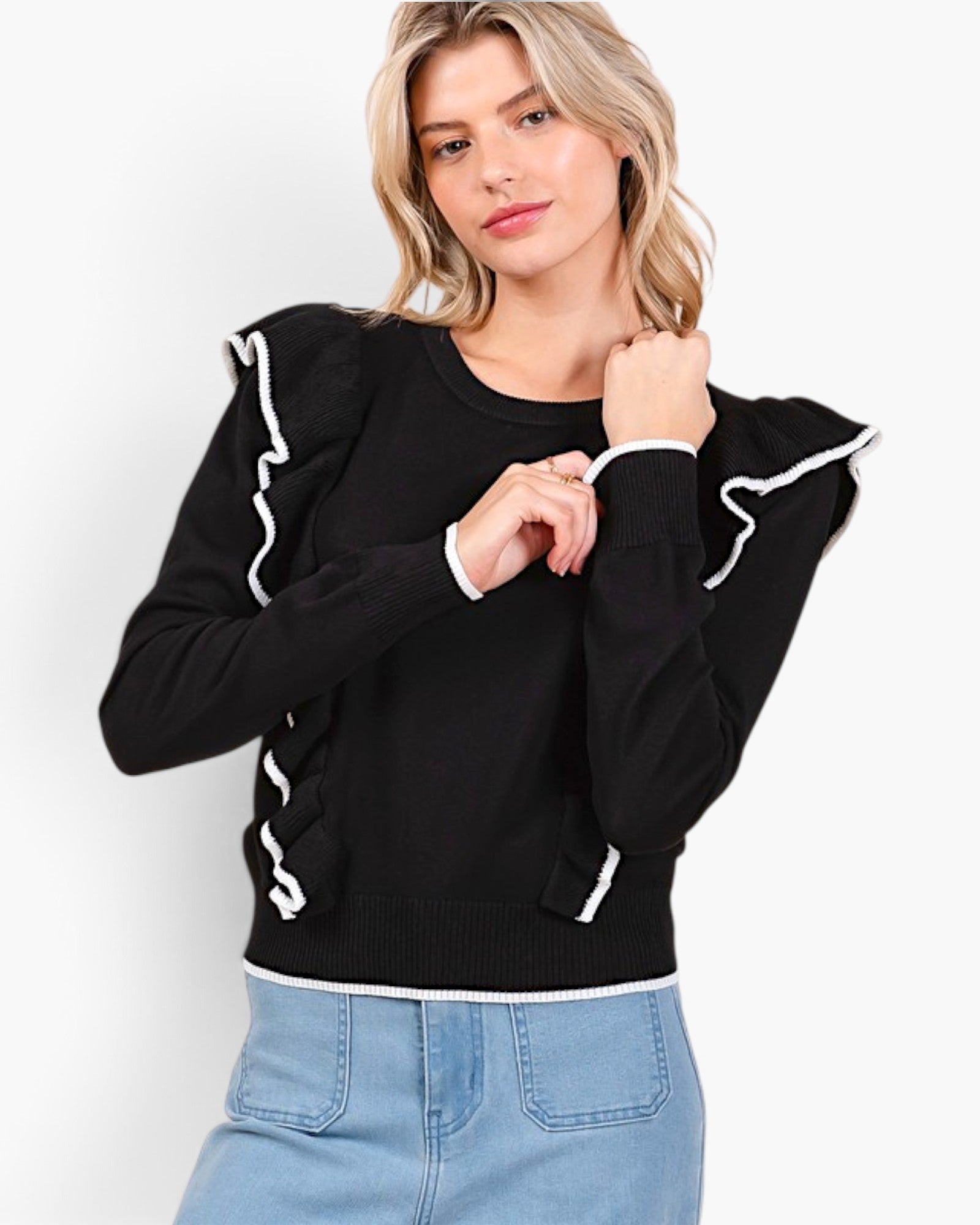 A woman wearing a black long-sleeve knit sweater with white contrast trim and ruffled shoulder details, paired with light blue jeans, against a plain white studio background.