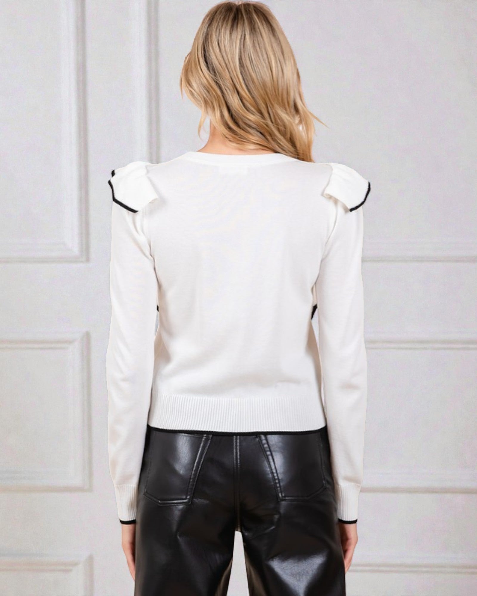 Person wearing a white blouse with black trim and black leather pants against a light-colored wall.