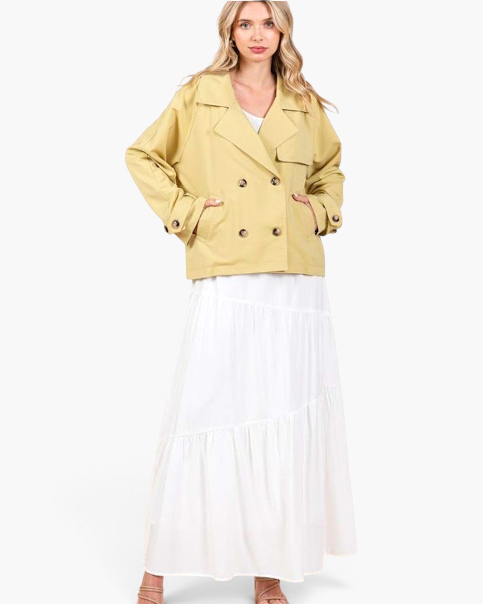 A woman modeling a cropped, double-breasted yellow jacket with brown buttons, worn over a white top and a long white tiered skirt, against a plain white studio background.
