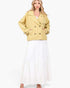 A woman modeling a cropped, double-breasted yellow jacket with brown buttons, worn over a white top and a long white tiered skirt, against a plain white studio background.