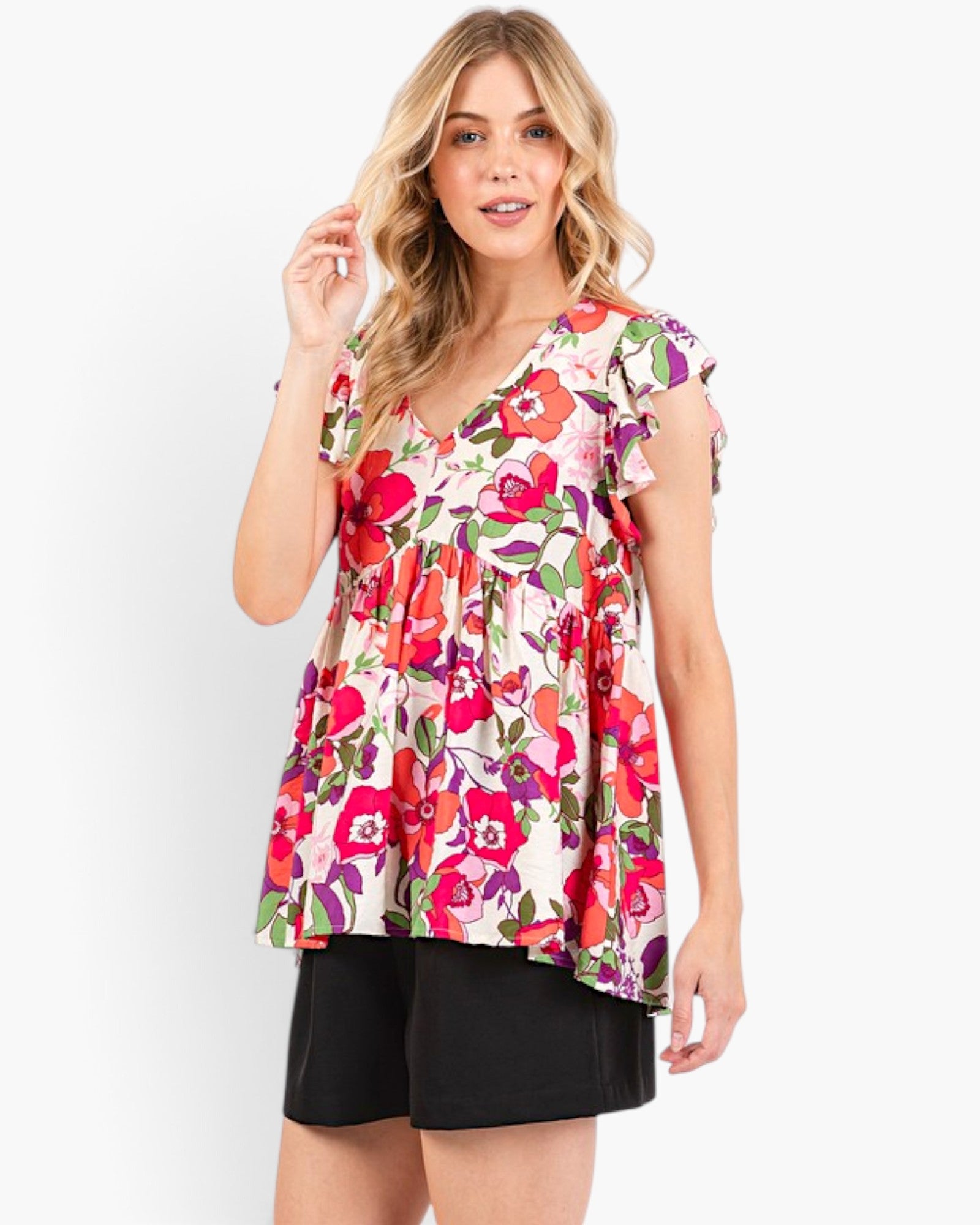 A woman modeling a short-sleeve V-neck peplum top with a vibrant red, pink, and green floral print on a cream background, paired with black shorts, against a plain white studio background.