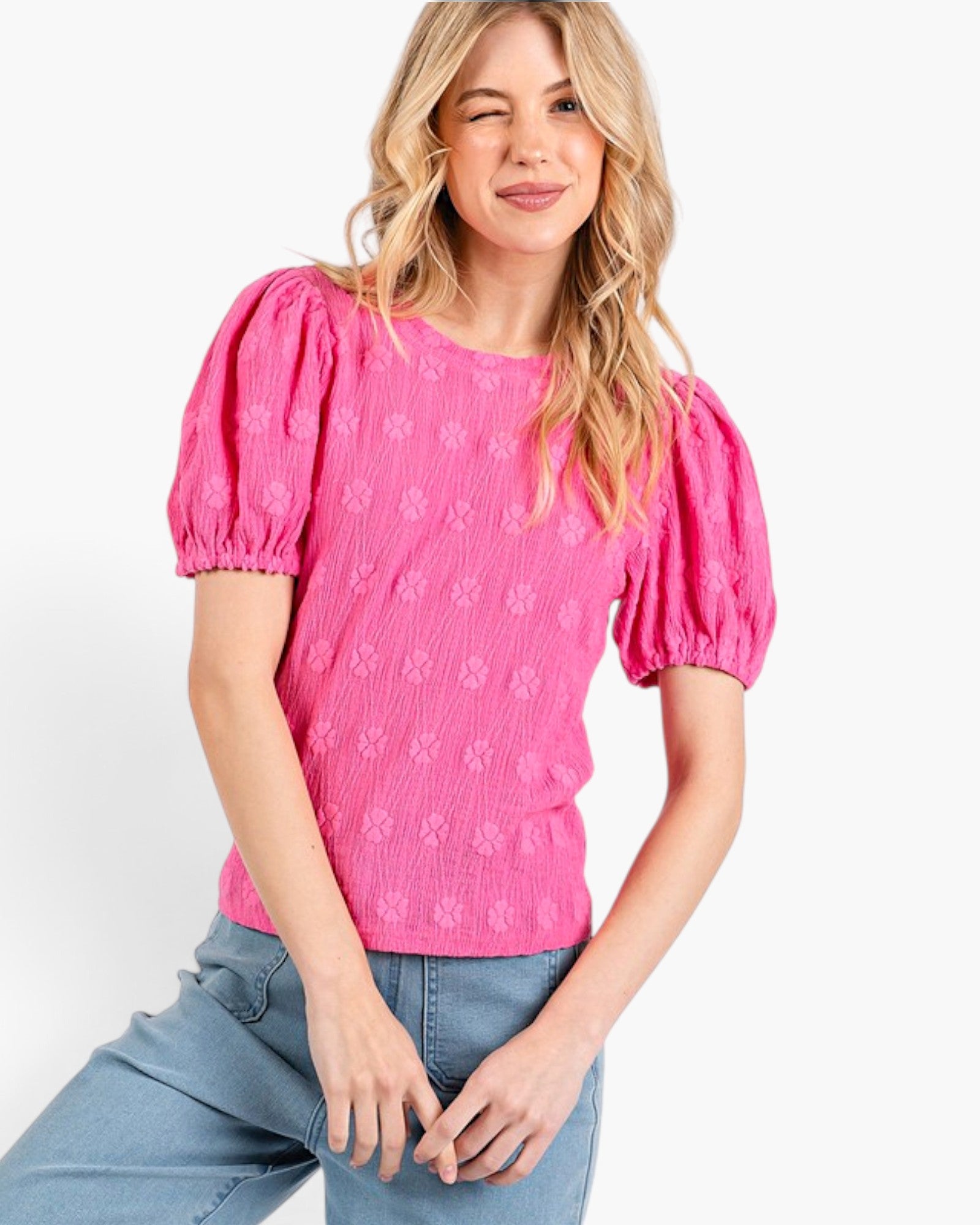 A woman wearing a bright pink textured short-sleeve top with puff sleeves and light blue jeans, posing against a clean white background.