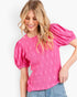 A woman wearing a bright pink short-sleeve top with textured puff sleeves and a subtle tonal floral pattern, paired with light blue jeans against a plain white background.