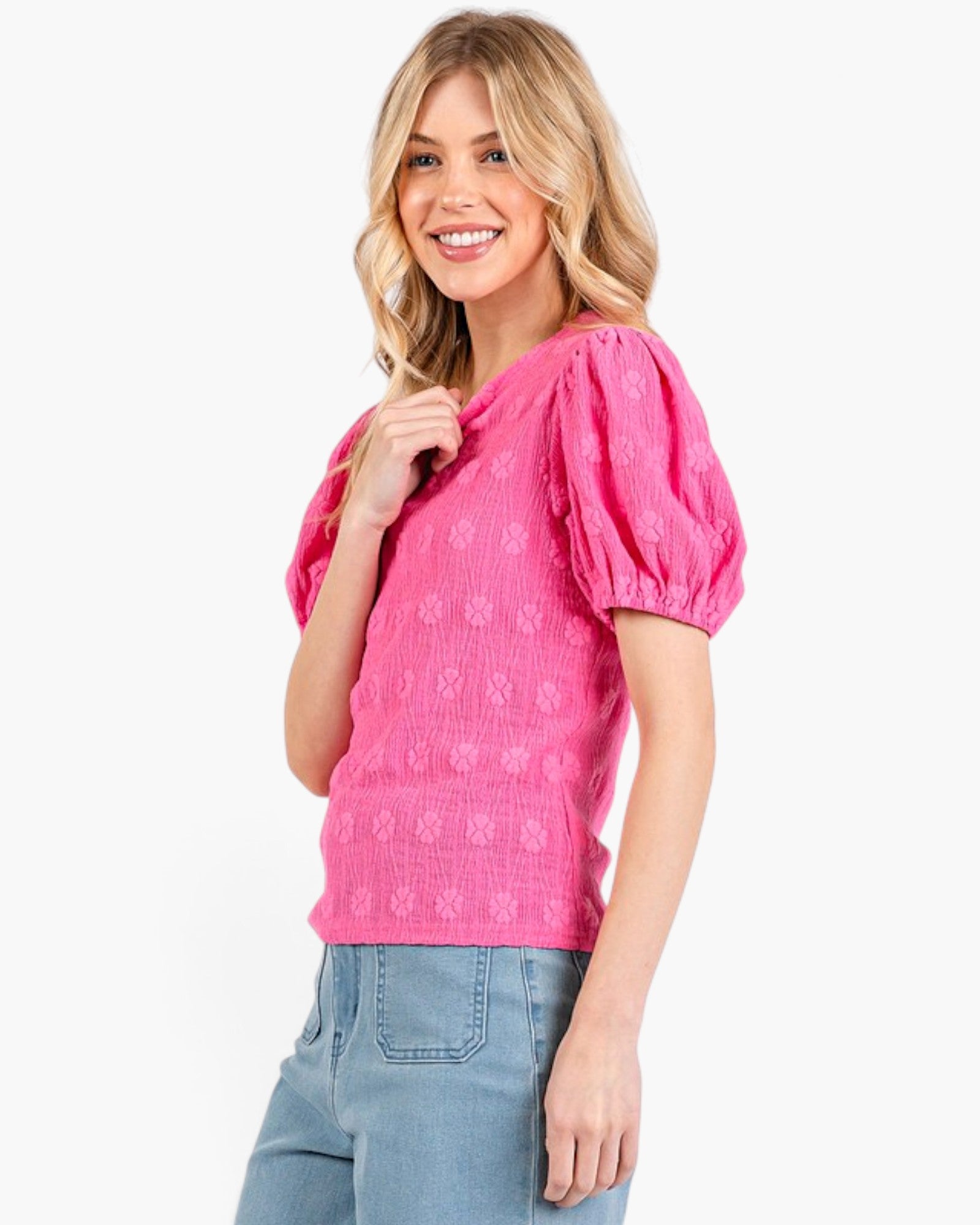A woman wearing a bright pink textured short-sleeve top with puff sleeves and light blue jeans, photographed against a clean white background.