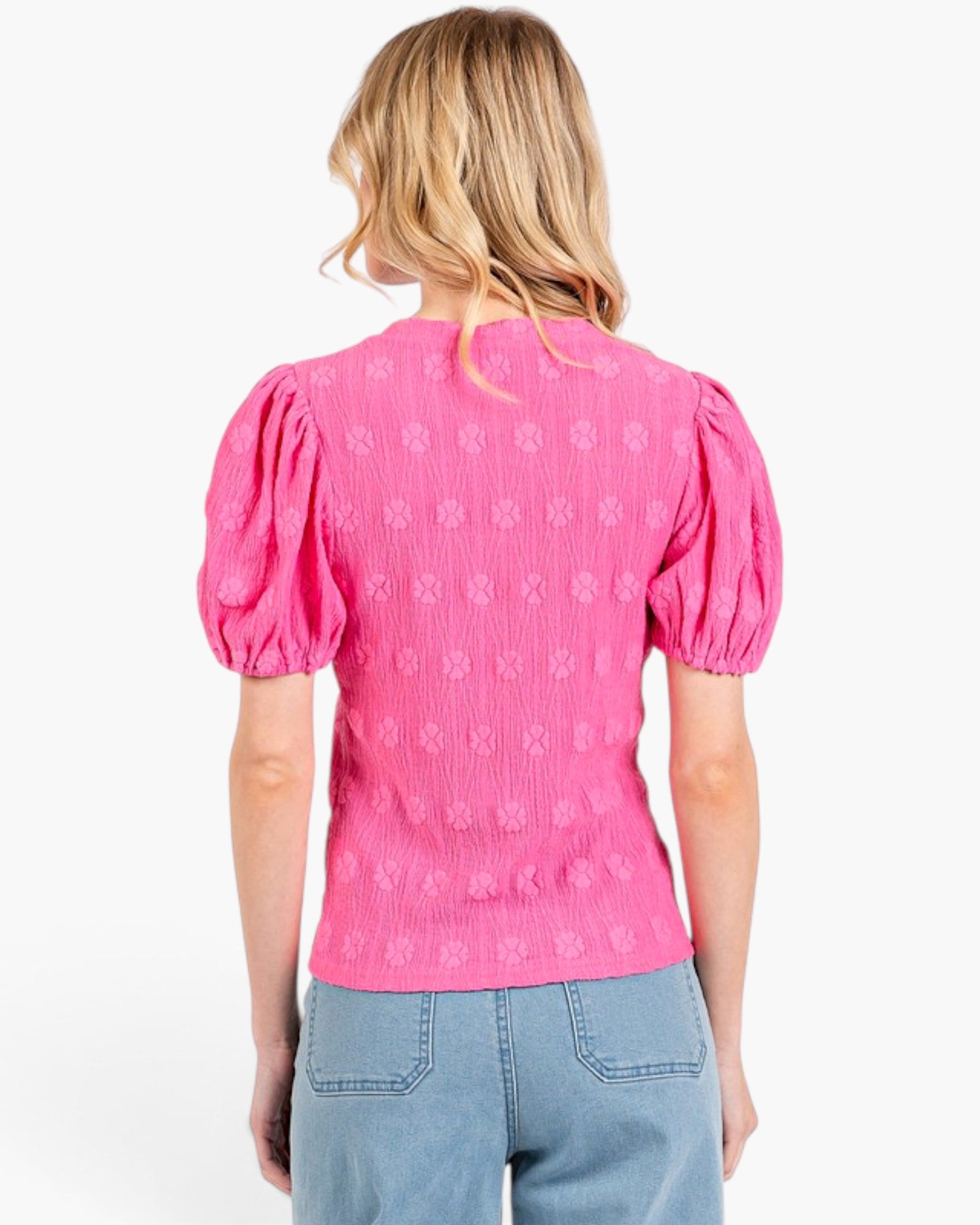 A woman viewed from the back wearing a bright pink textured short-sleeve top with puffed sleeves and light blue jeans, against a plain white background.