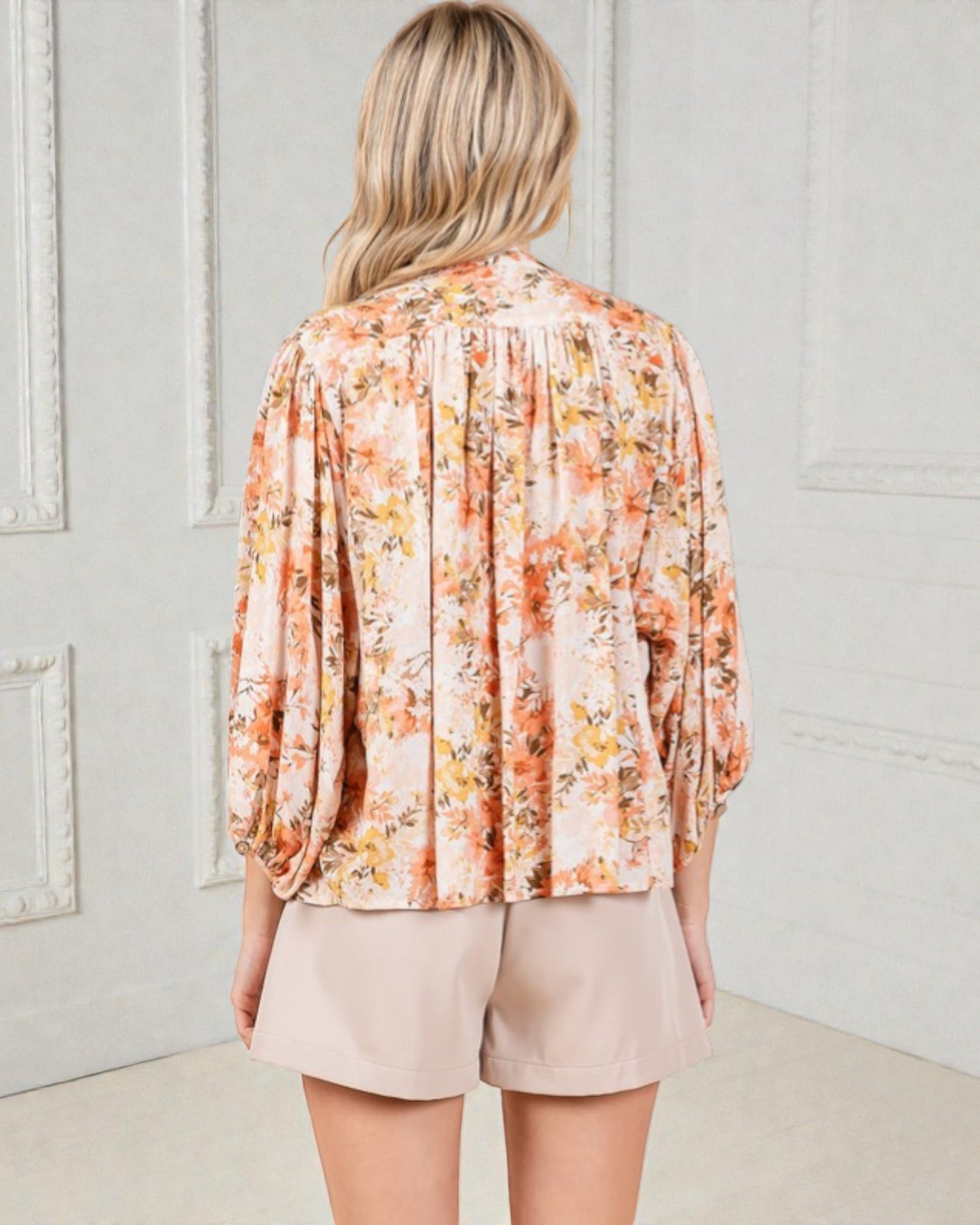 Woman wearing a floral blouse and beige shorts against a light gray wall.