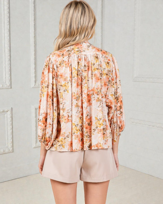 Woman wearing a floral blouse and beige shorts against a light gray wall.