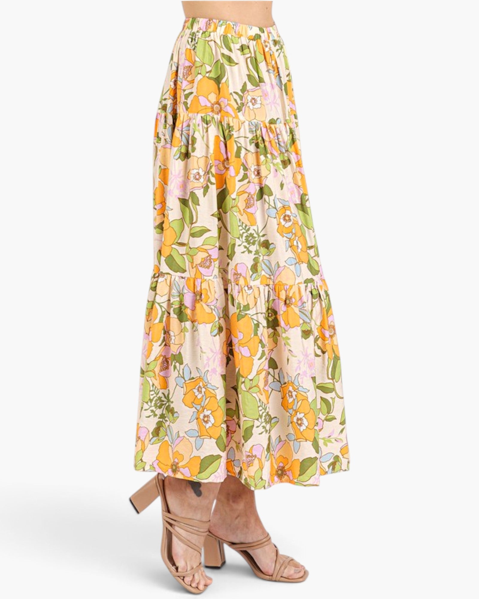 A model is shown from the waist down wearing a high-waisted, tiered maxi skirt with a vibrant floral print in orange, yellow, green, and pink on a cream background, paired with beige strappy high-heeled sandals against a light gray studio background.
