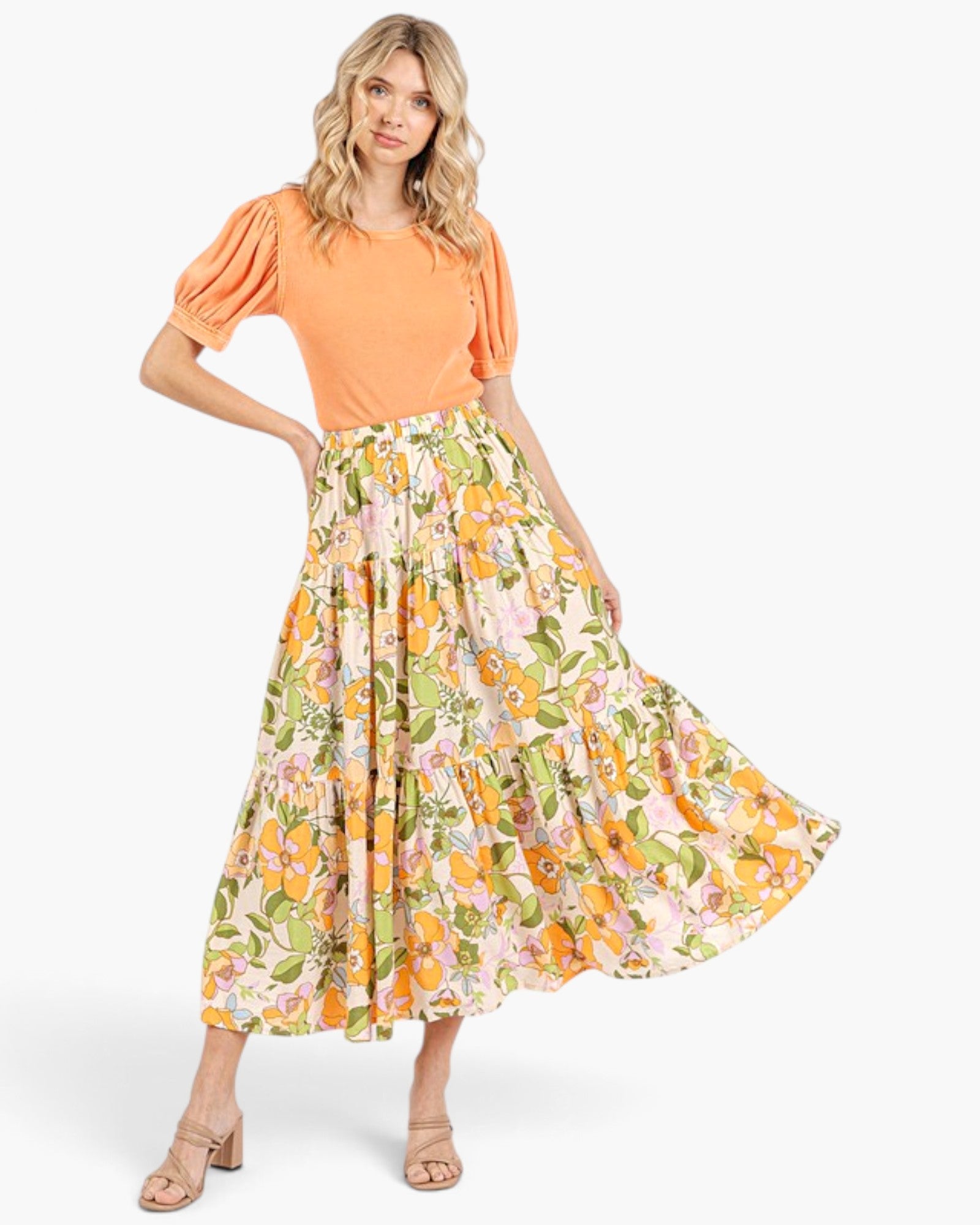 A woman modeling a two-piece outfit consisting of a short-sleeve orange puff-sleeve top and a high-waisted, tiered maxi skirt with a vibrant orange, yellow, and green floral print on a cream background.