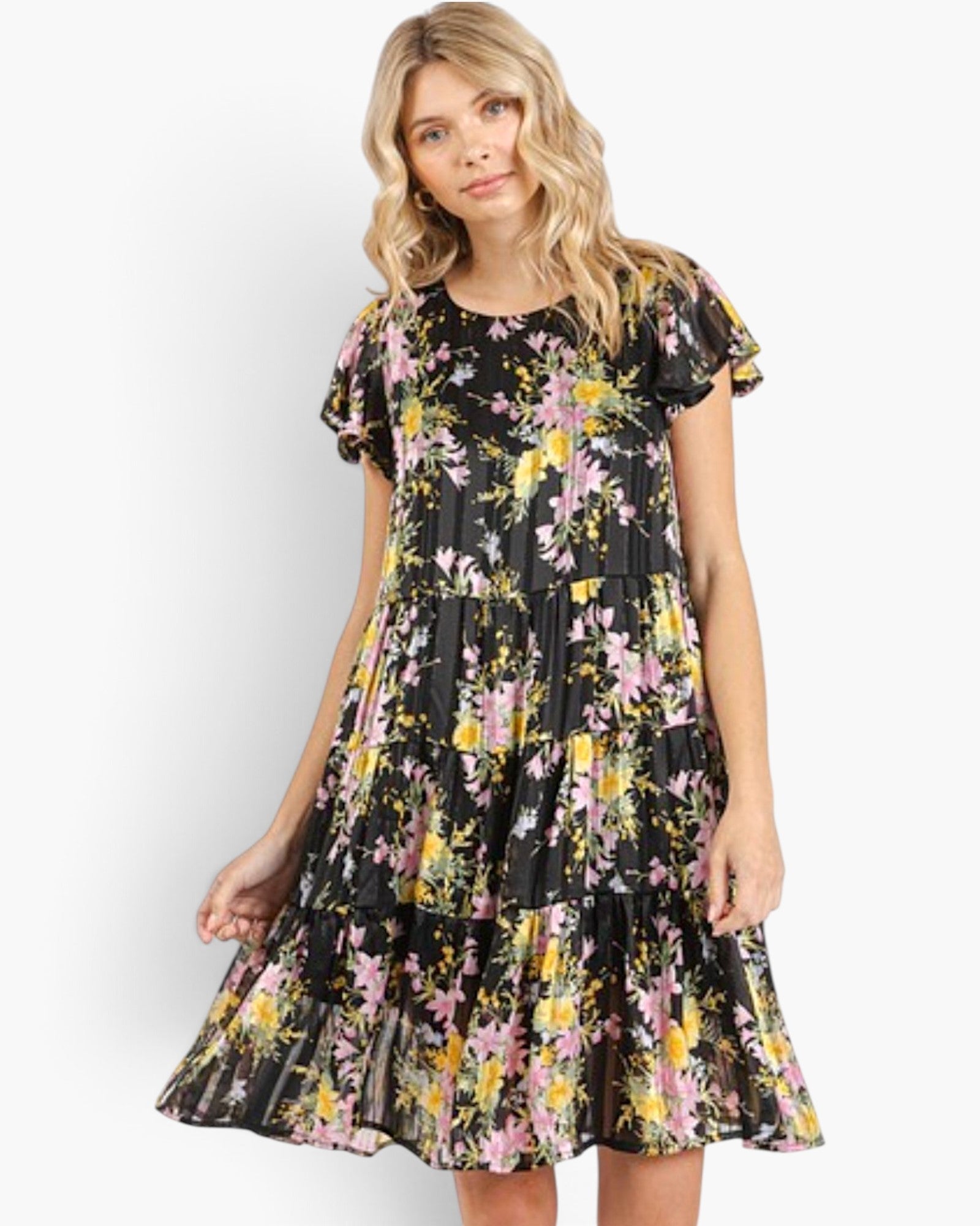 A woman modeling a tiered black dress with a vibrant pink, yellow, and green floral print against a plain light gray background.