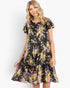 A woman modeling a tiered black dress with a vibrant pink, yellow, and green floral print against a plain light gray background.