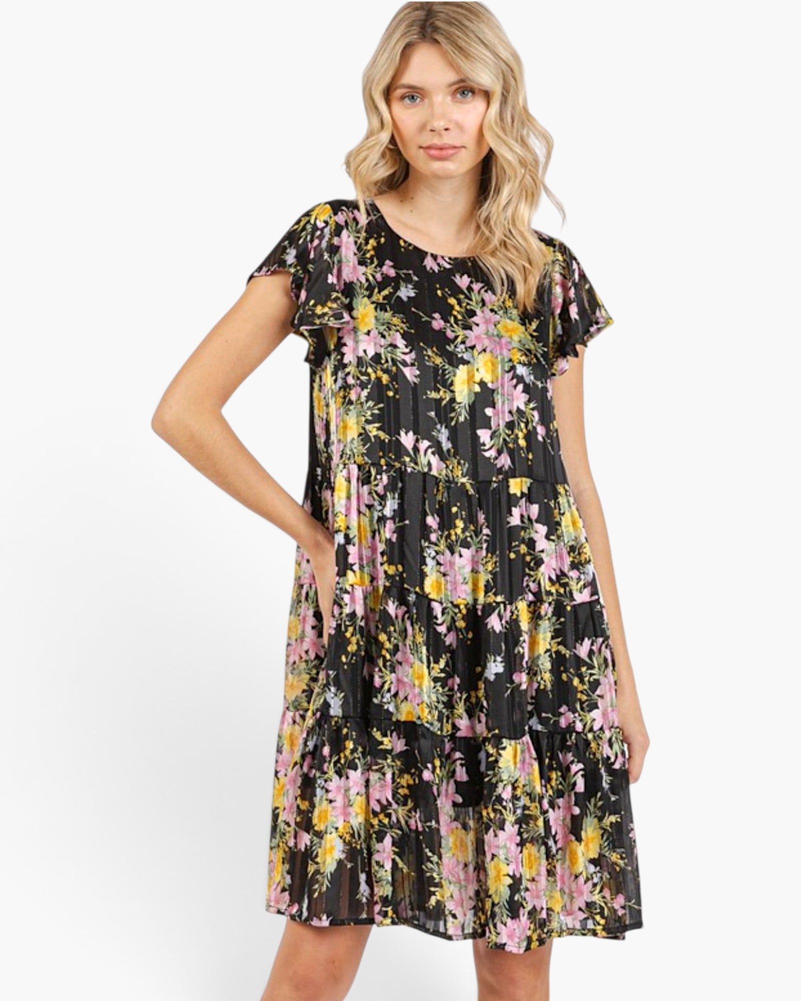 A woman modeling a short-sleeve tiered dress with a black background and a colorful floral print in pink, yellow, and white, set against a plain white studio background.