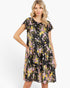 A woman modeling a short-sleeve tiered dress with a black background and a colorful floral print in pink, yellow, and white, set against a plain white studio background.
