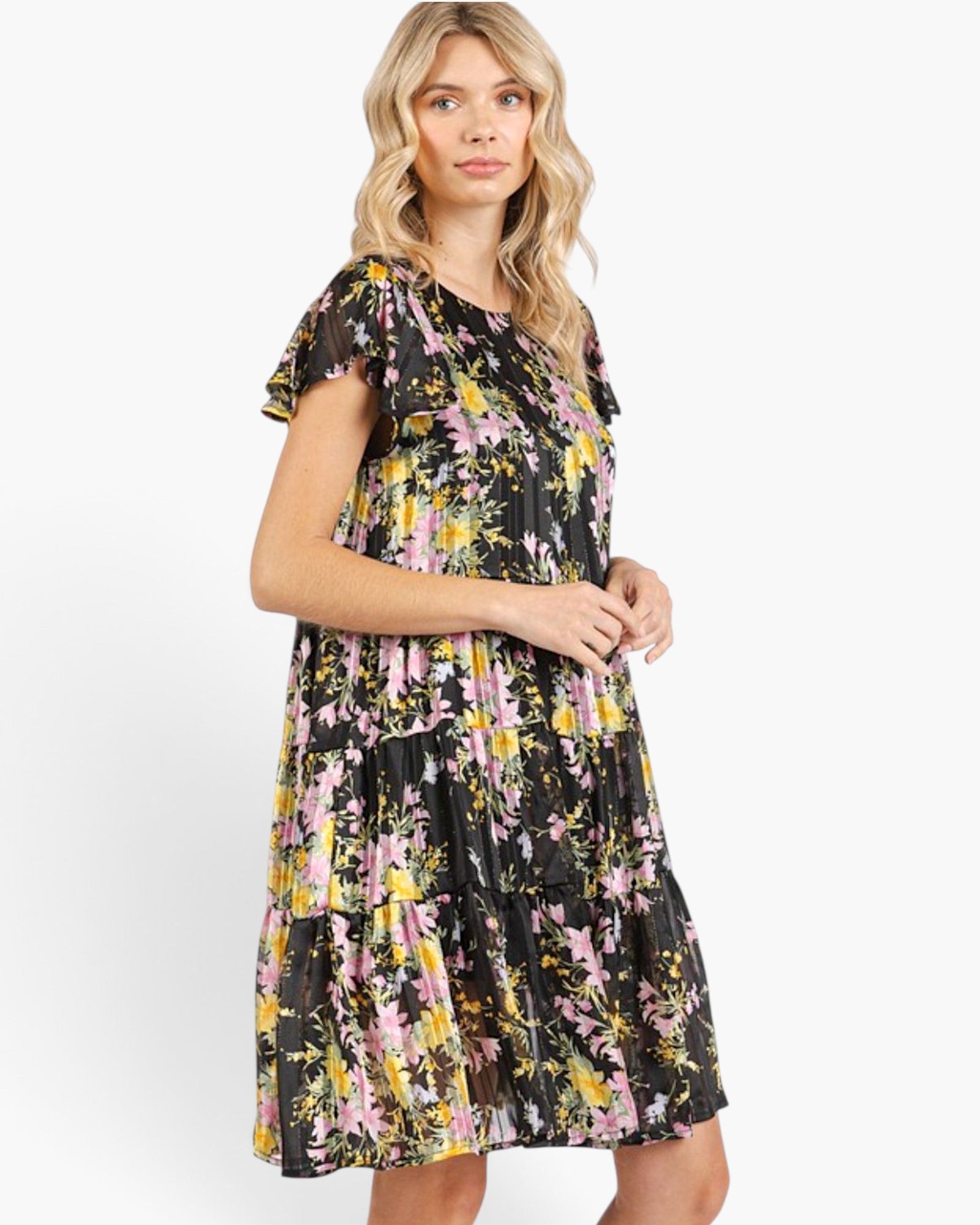 A woman modeling a tiered black midi dress with a vibrant pink, yellow, and white floral print against a plain white studio background.