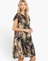 A woman modeling a tiered black midi dress with a vibrant pink, yellow, and white floral print against a plain white studio background.