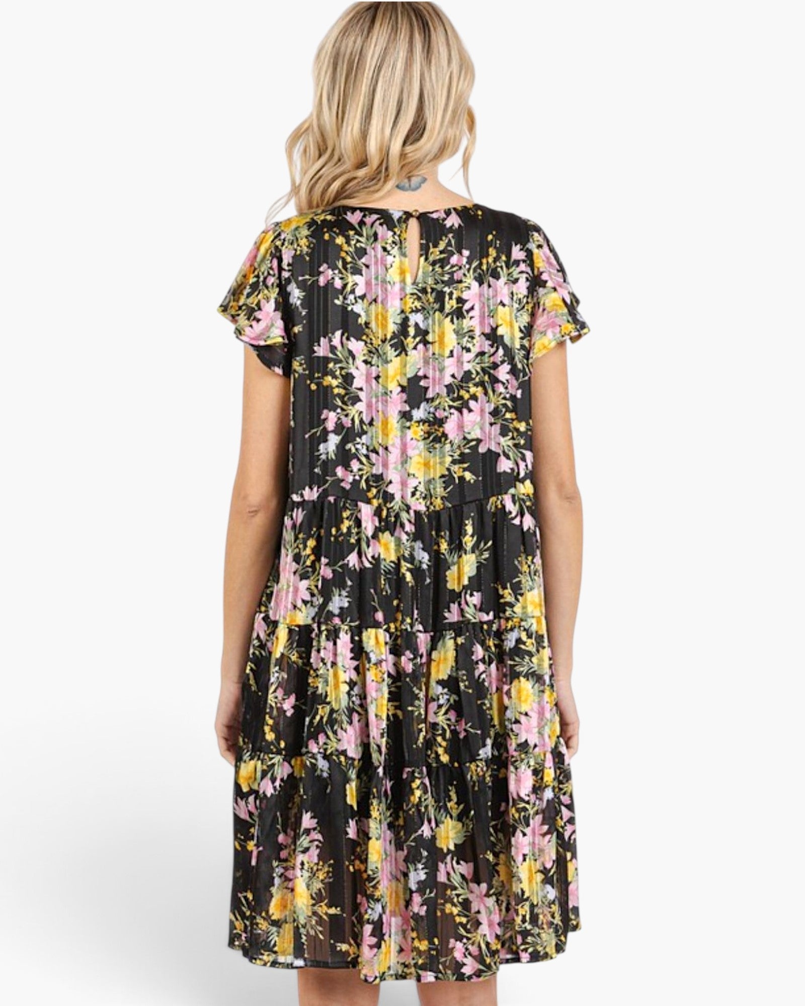 A woman viewed from the back wearing a short-sleeve pleated dress with a black base and a pink, yellow, and white floral print, set against a plain light gray studio background.