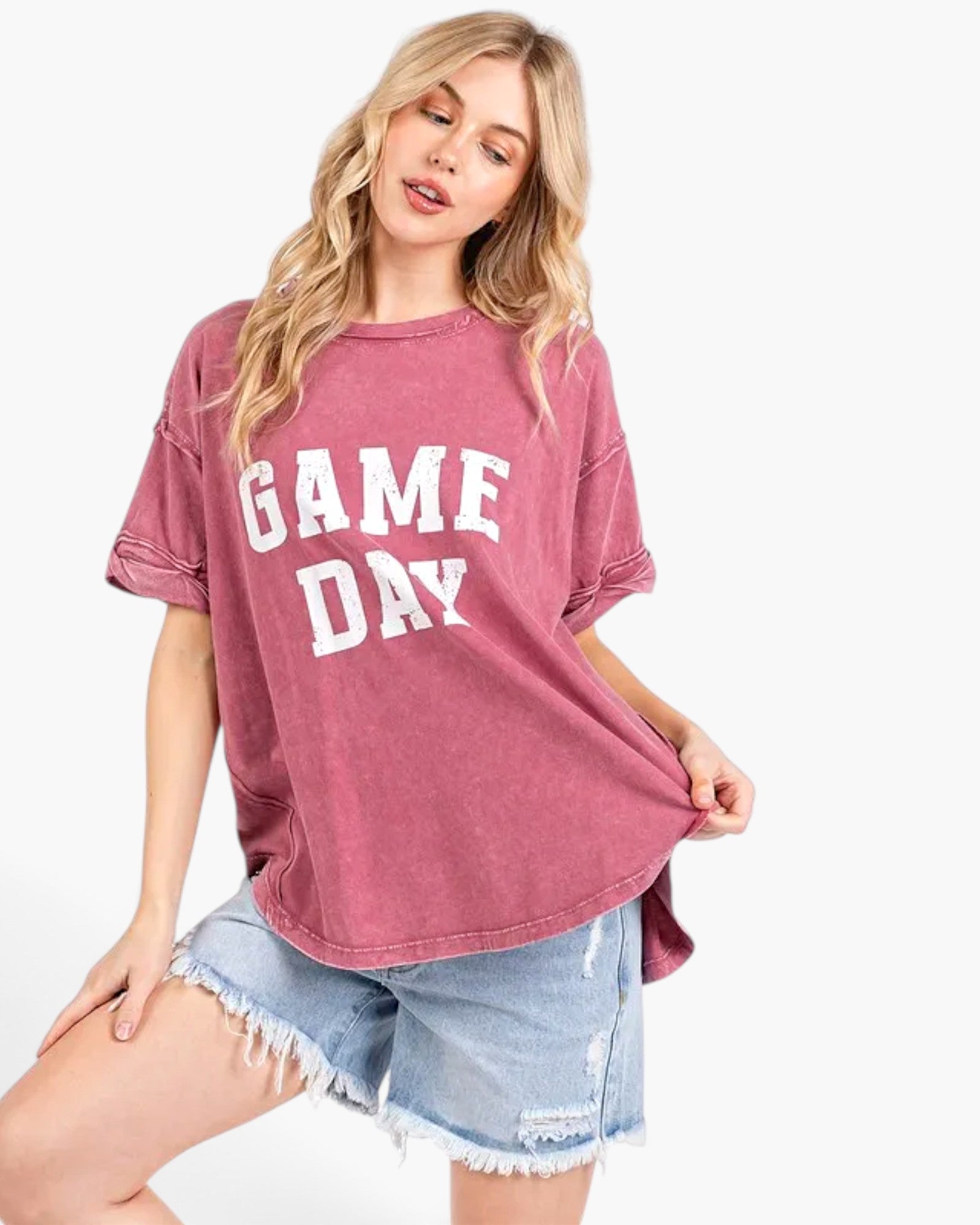 A woman modeling a washed pink short-sleeve t-shirt with 'GAME DAY' printed in white block letters across the front, paired with light blue denim shorts against a plain white background.