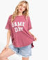 A woman modeling a washed pink short-sleeve t-shirt with 'GAME DAY' printed in white block letters across the front, paired with light blue denim shorts against a plain white background.