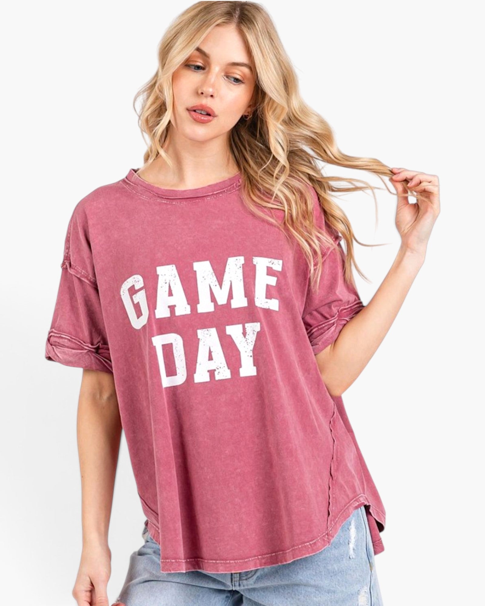 A woman wearing a washed dusty rose short-sleeve t-shirt with 'GAME DAY' printed in white block letters on the front, paired with light blue jeans against a plain white background.
