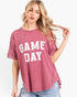 A woman wearing a washed dusty rose short-sleeve t-shirt with 'GAME DAY' printed in white block letters on the front, paired with light blue jeans against a plain white background.