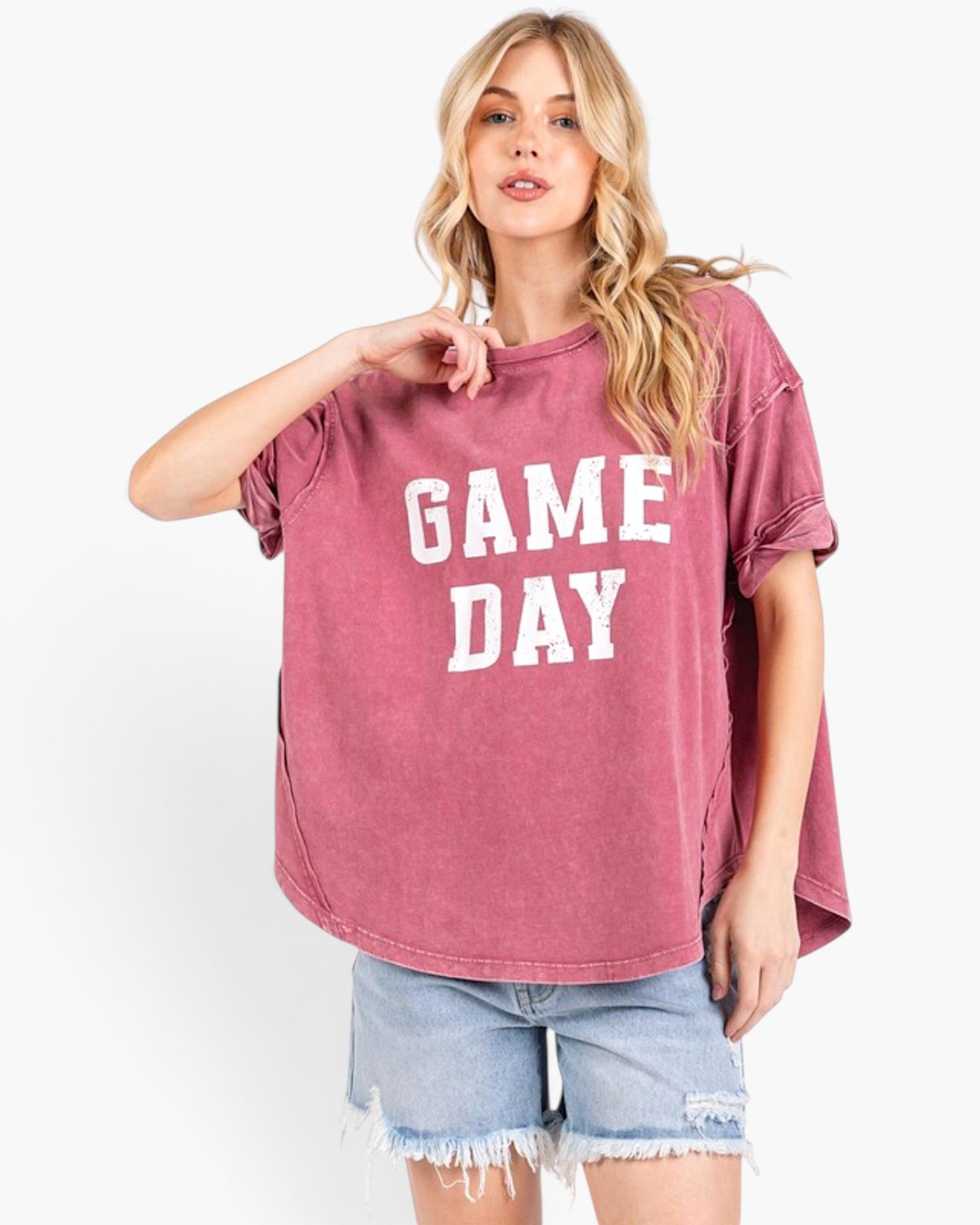 A woman modeling a washed pink short-sleeve t-shirt with 'GAME DAY' printed in white block letters across the front, paired with light blue denim shorts against a white studio background.