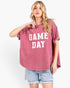A woman modeling a washed pink short-sleeve t-shirt with 'GAME DAY' printed in white block letters across the front, paired with light blue denim shorts against a white studio background.