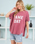 Woman wearing a 'GAME DAY' t-shirt in a casual setting with a plant and shelves in the background.