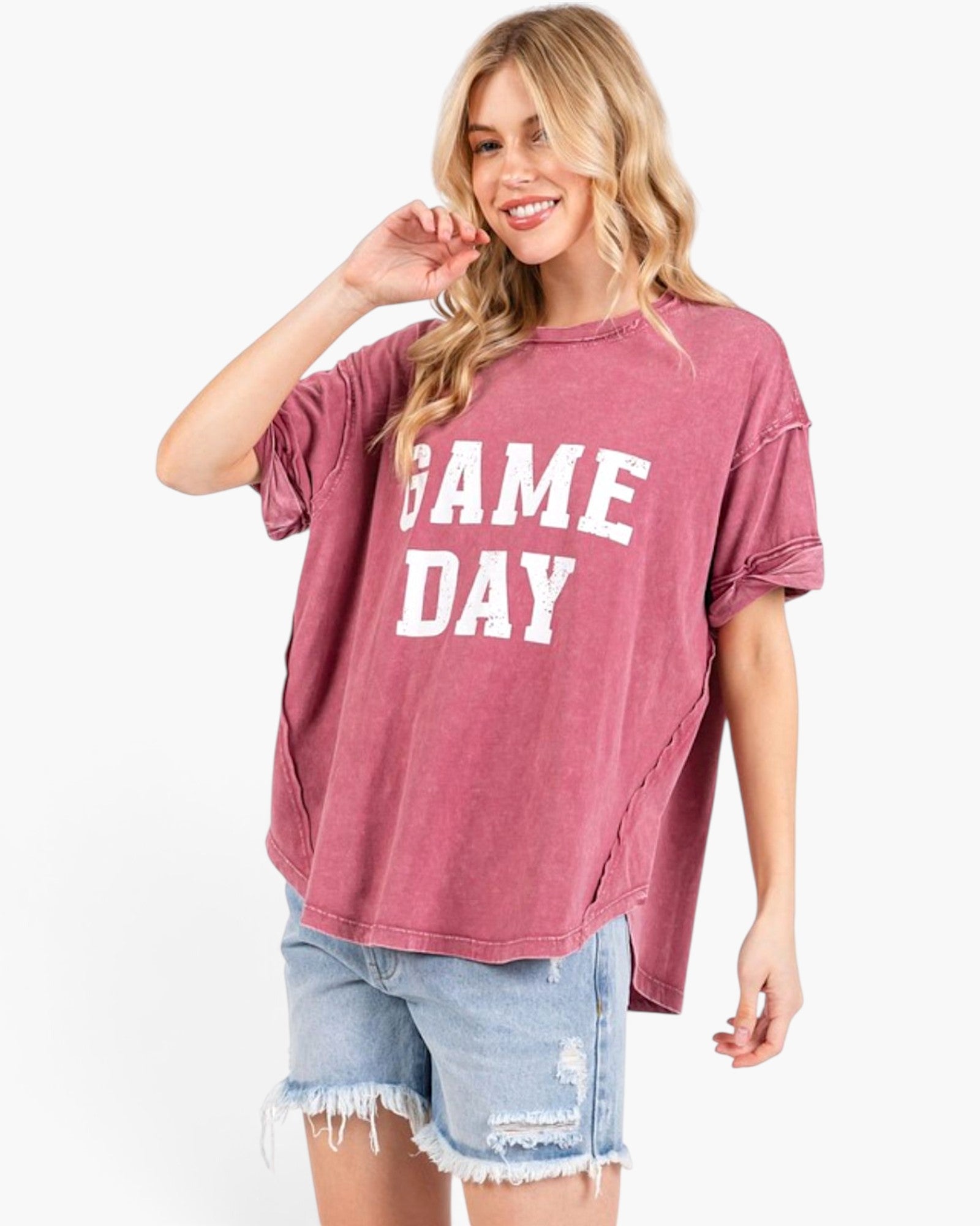 A woman modeling a washed dusty rose short-sleeve t-shirt with 'GAME DAY' printed in white block letters across the front, paired with light blue distressed denim shorts against a plain white background.