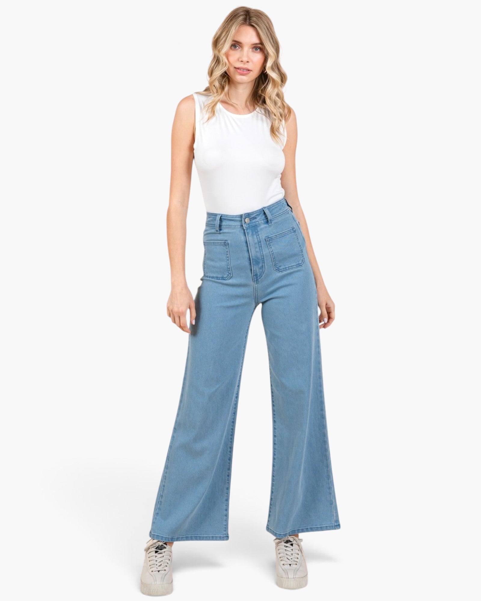 A woman modeling a pair of high-waisted, wide-leg light blue denim jeans with a white sleeveless top and white sneakers, set against a plain white studio background.
