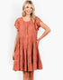 A woman modeling a terracotta-colored tiered dress with ruffled short sleeves, button details, and embroidered horizontal bands, set against a plain white studio background.