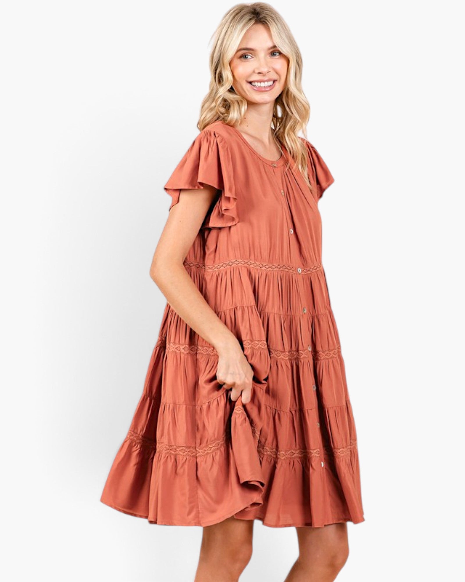 A woman modeling a terracotta-colored tiered dress with ruffled sleeves and embroidered details, set against a plain white studio background.