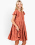 A woman modeling a terracotta-colored tiered dress with ruffled sleeves and embroidered details, set against a plain white studio background.