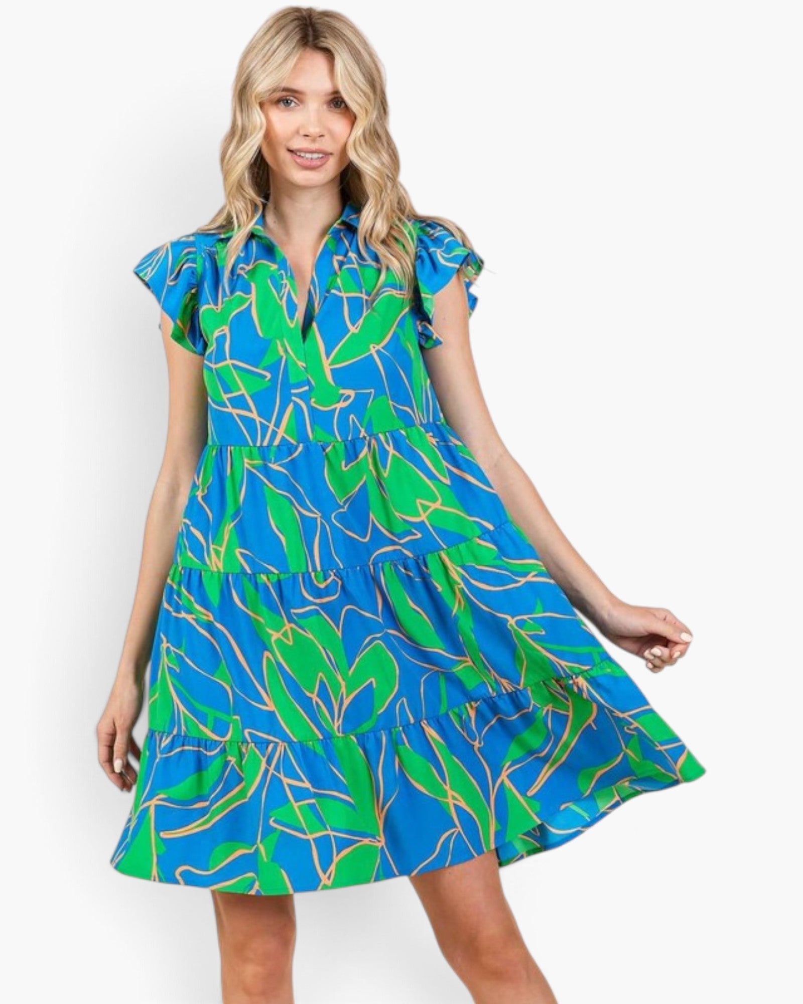 A woman modeling a short-sleeve tiered dress with a vibrant blue and green tropical leaf print outlined in yellow, set against a plain light gray background.