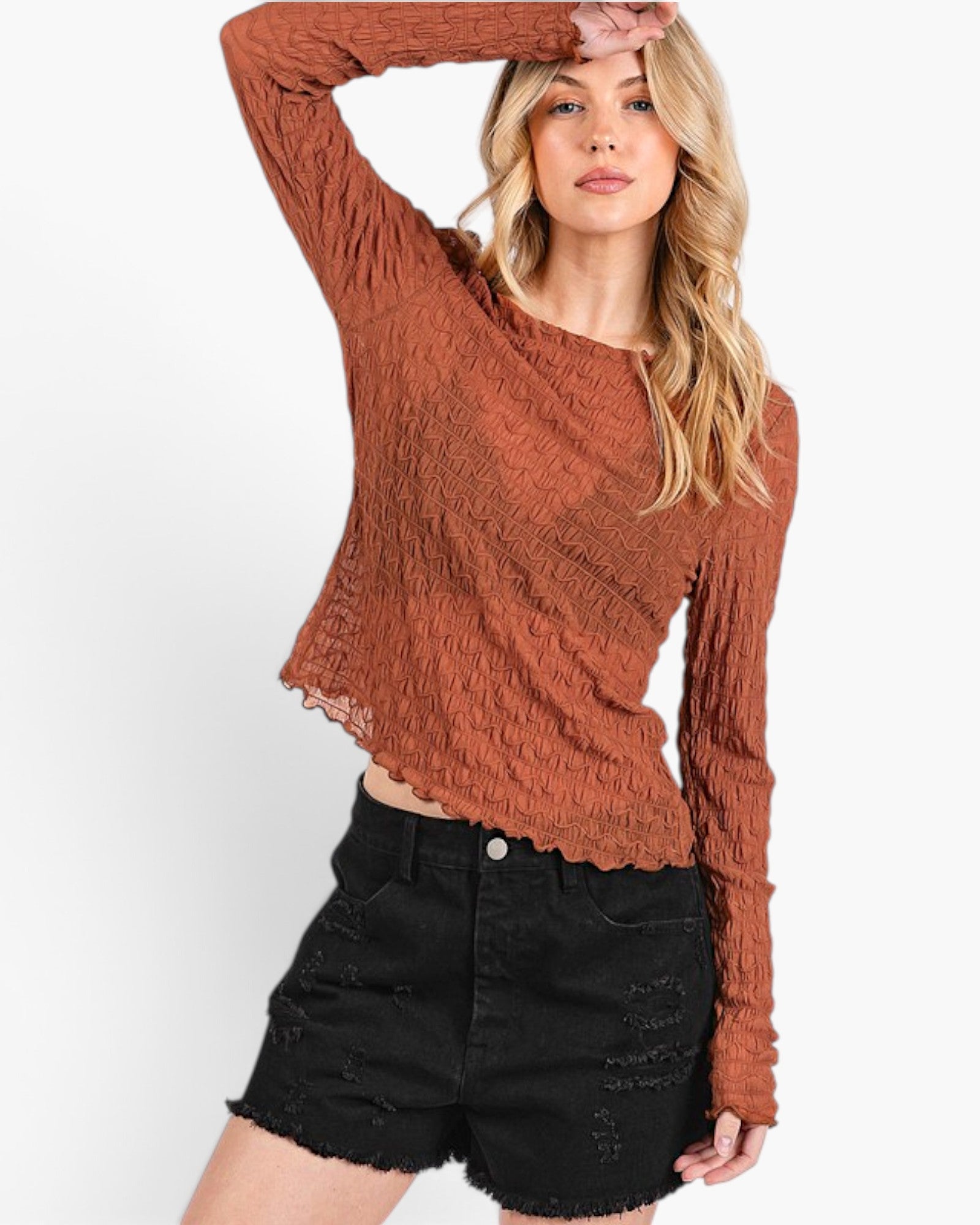 A woman modeling a textured, long-sleeve cropped top in a rust orange color, paired with black distressed denim shorts, against a plain white background.