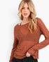 A woman wearing a textured, long-sleeve rust-colored top with a smocked chest panel and a relaxed fit, paired with black pants, against a plain white background.