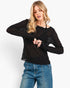 A woman wearing a black long-sleeve textured knit top with lace details and blue jeans, photographed against a clean white studio background.