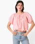 A woman modeling a light pink short-sleeve crop top with puff sleeves and a tie neckline, paired with light-wash blue jeans against a plain white background.