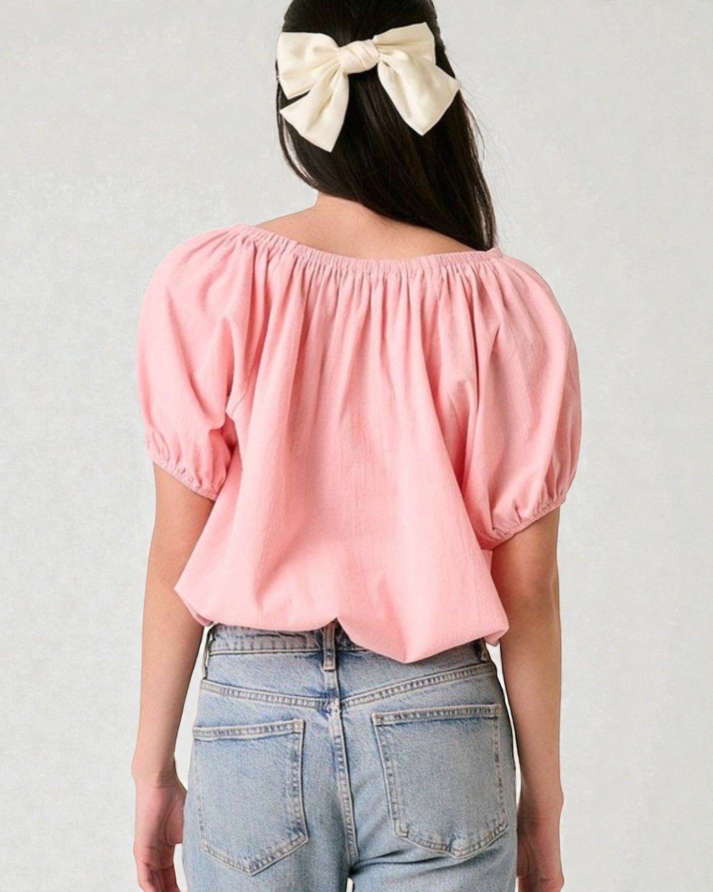 Person wearing a pink off-shoulder top and blue jeans with a white bow in hair on a light gray background
