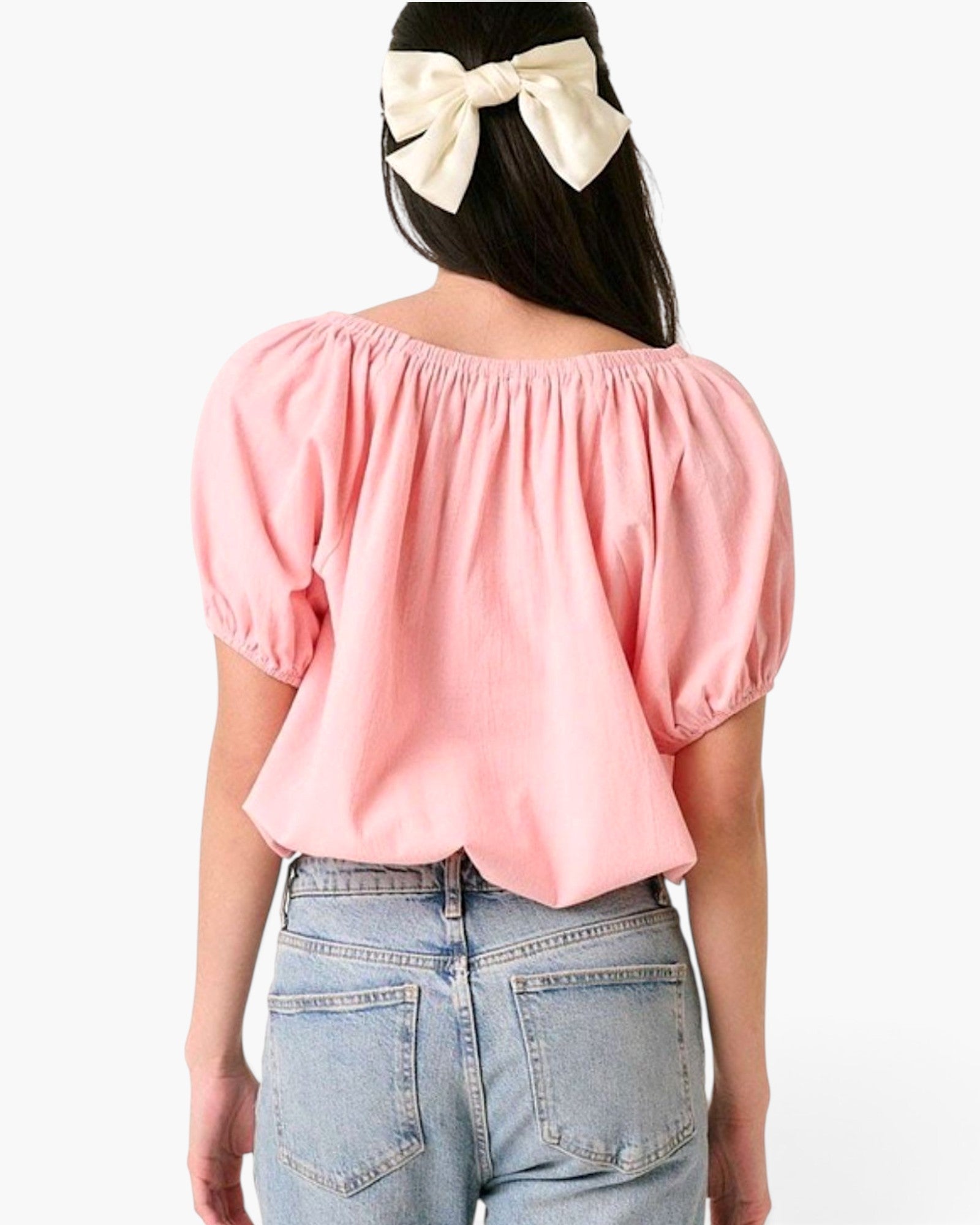 A woman shown from the back wearing a light pink puff-sleeve top and light blue jeans, with a cream bow in her dark hair, against a plain white background.