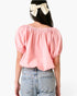 A woman shown from the back wearing a light pink puff-sleeve top and light blue jeans, with a cream bow in her dark hair, against a plain white background.