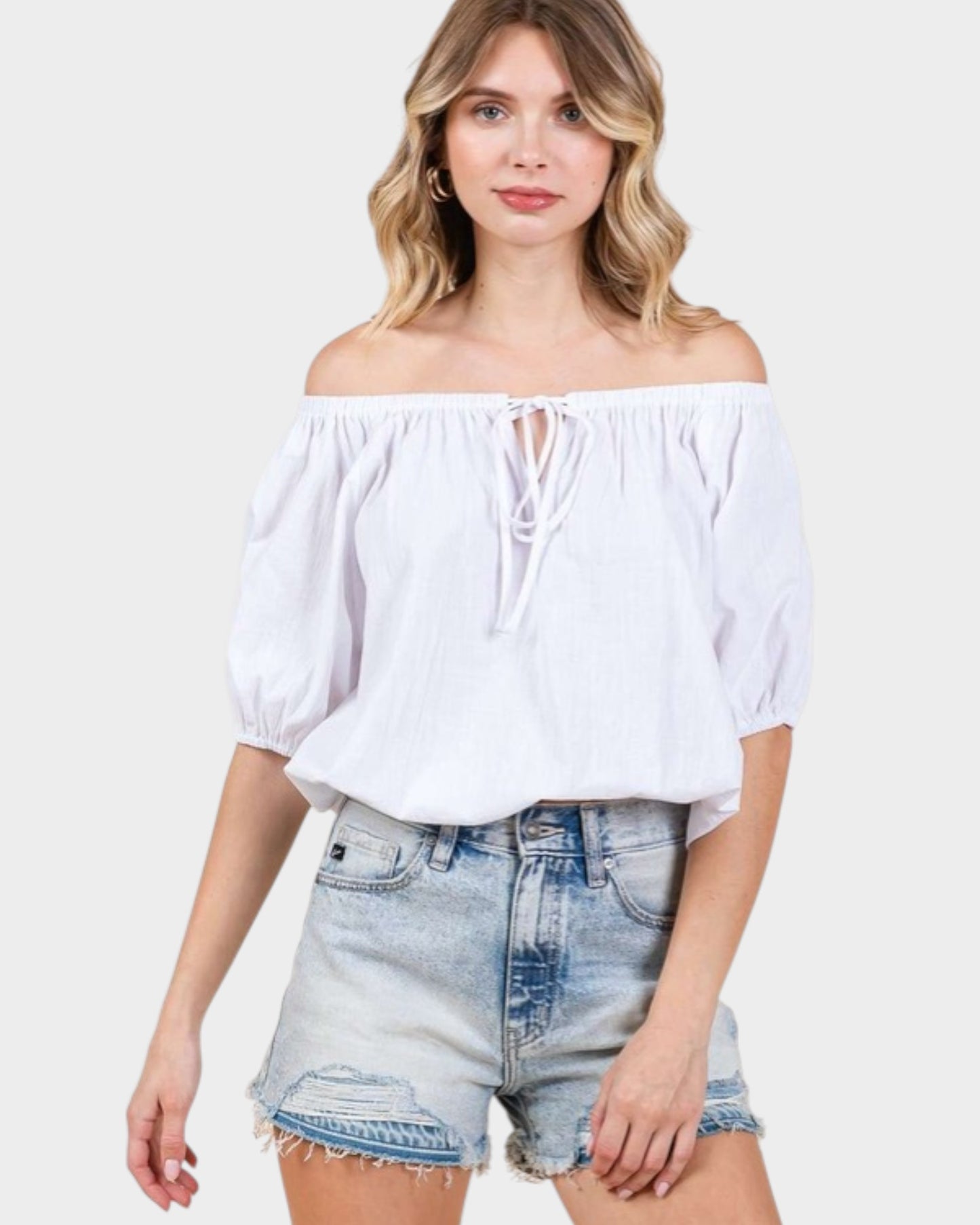 Woman wearing a white off-shoulder top and denim shorts on a plain background