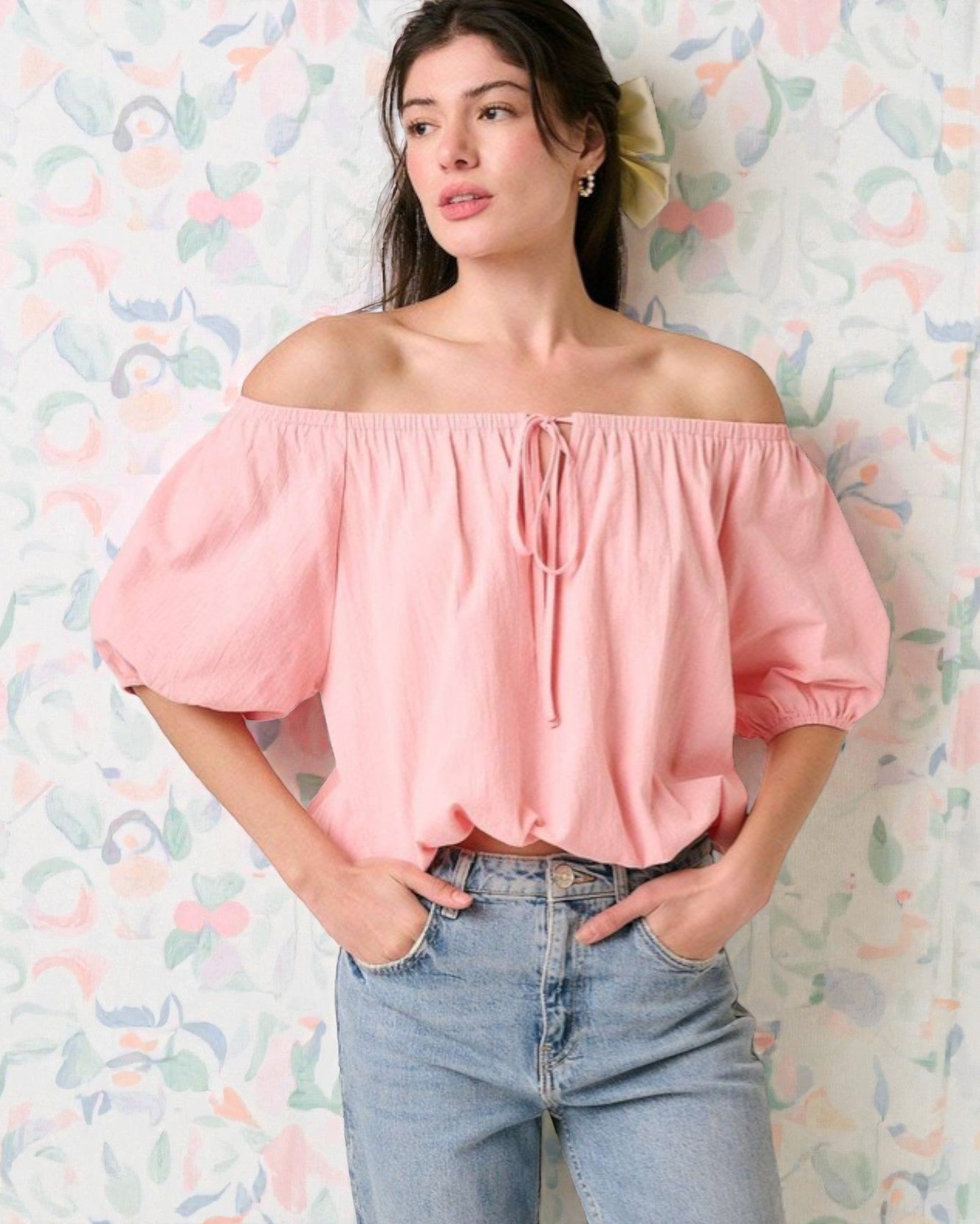 Woman wearing a pink off-shoulder top and blue jeans against a colorful abstract background