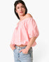 A woman wearing a light pink off-the-shoulder puff-sleeve crop top and light-wash blue jeans, standing against a plain white studio background.