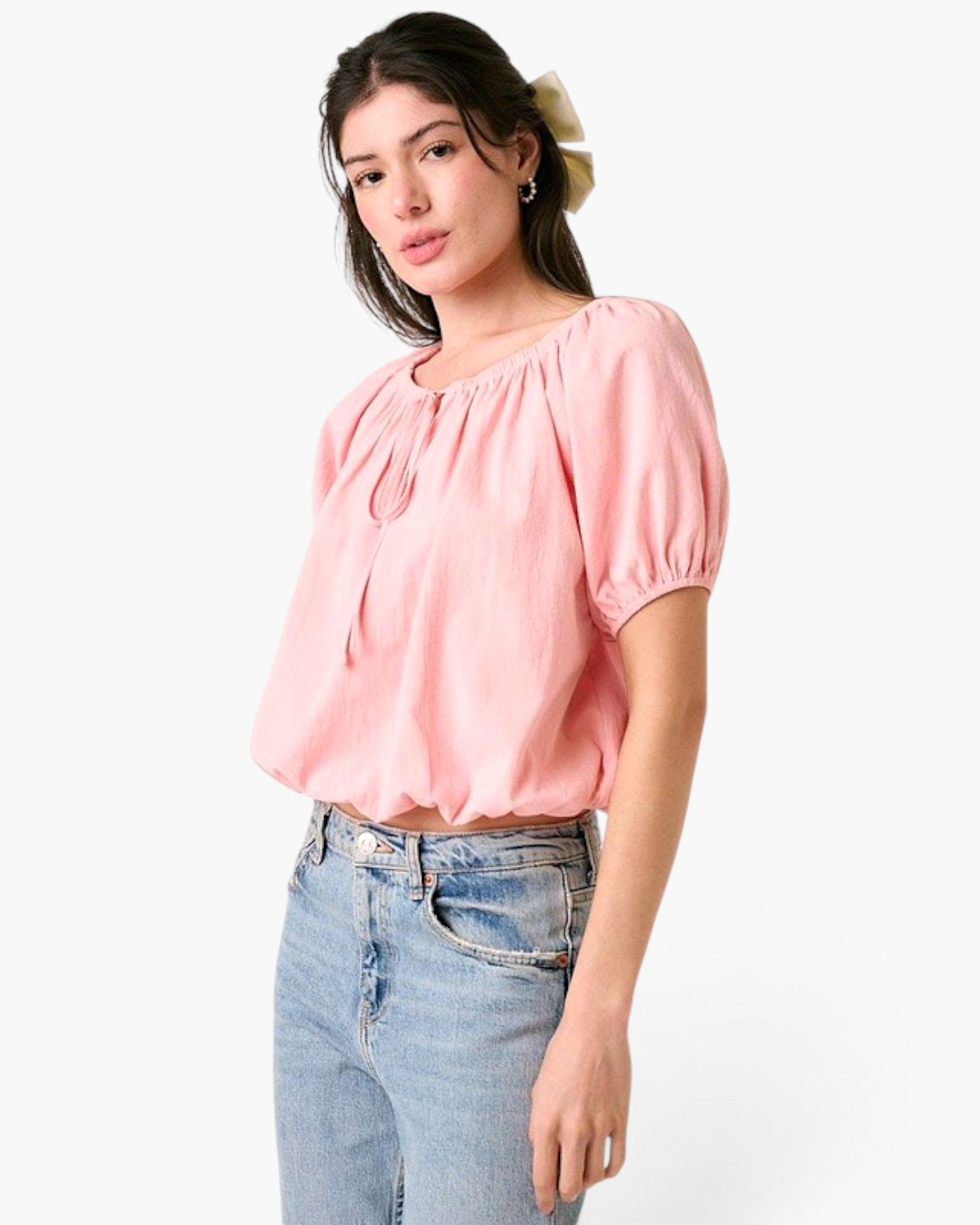 A woman wearing a light pink short-sleeve blouse with a gathered, puffed neckline and tie detail, paired with light-wash blue jeans, against a plain white background.