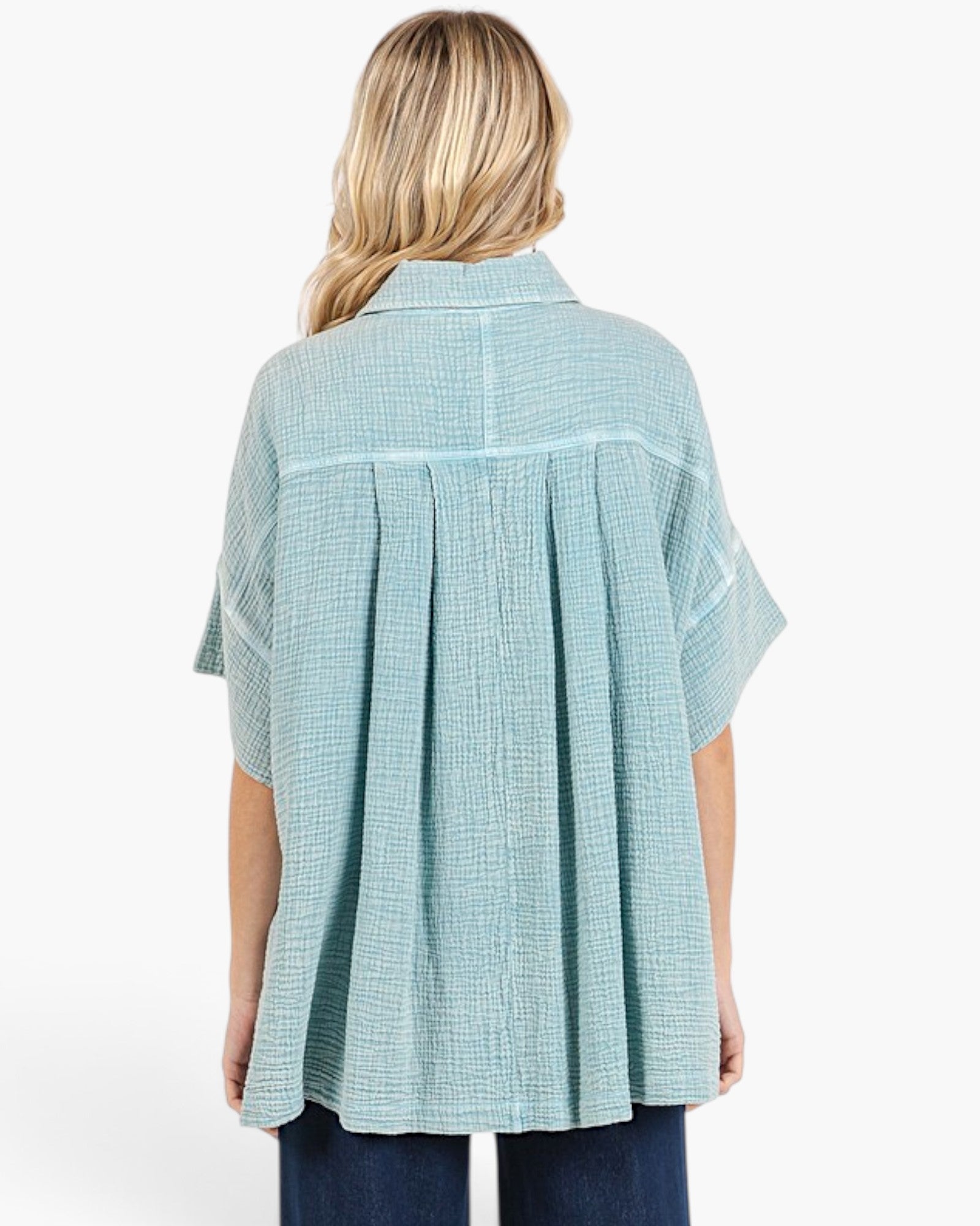 A woman viewed from the back wearing a light blue-green short-sleeve button-up shirt with a subtle grid pattern and pleated back detail, paired with dark blue jeans against a plain white background.
