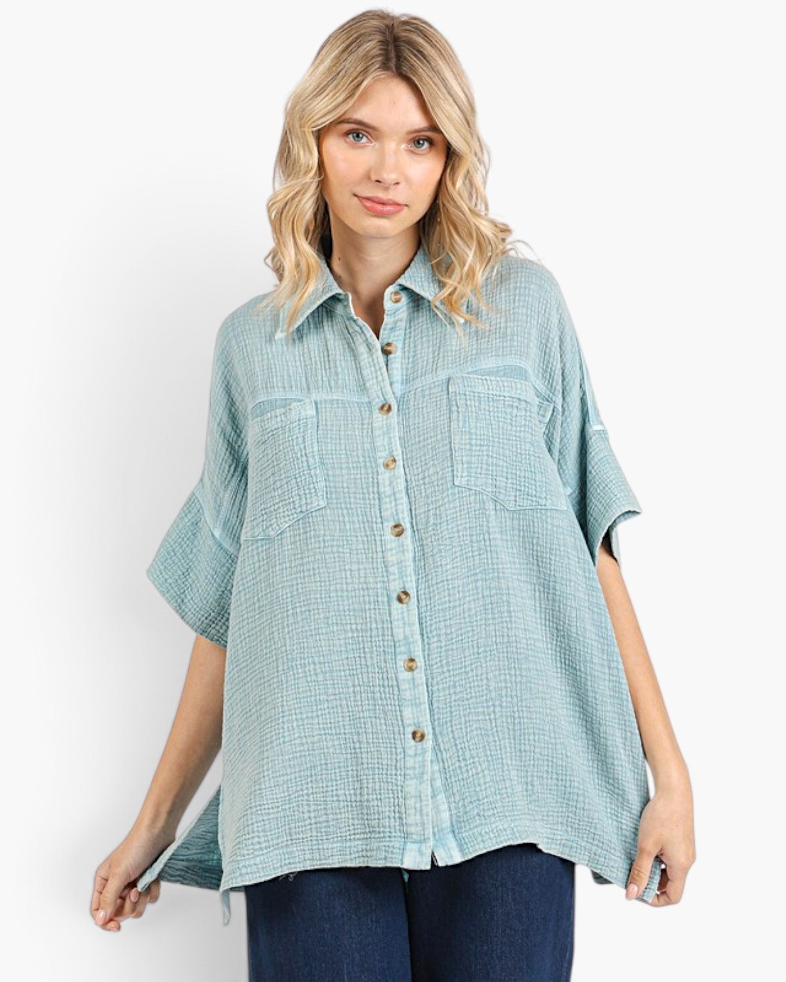 A woman modeling a light blue-green, short-sleeve button-up shirt with a subtle grid pattern, featuring two chest pockets and brown buttons, paired with dark blue jeans against a plain white background.