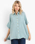 A woman modeling a light blue-green, short-sleeve button-up shirt with a subtle grid pattern, featuring two chest pockets and brown buttons, paired with dark blue jeans against a plain white background.