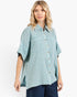 A woman wearing a light blue-green textured short-sleeve button-up shirt with dark blue jeans, photographed against a clean white studio background.